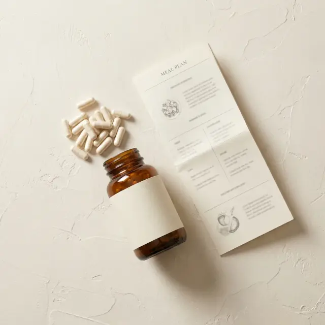 An amber glass bottle with a blank label, loose capsules and a printed plan, laid out on a pale plaster surface.