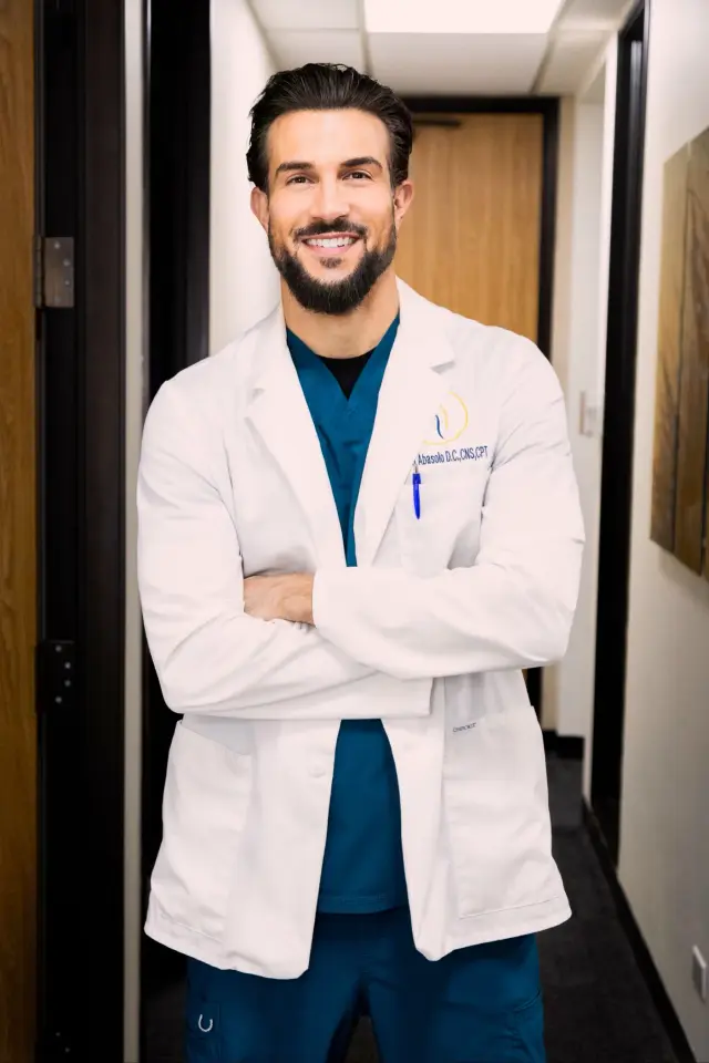 Bryan Abasolo, DC, arms folded, in a white coat embroidered with his name and post-nominals over teal scrubs.